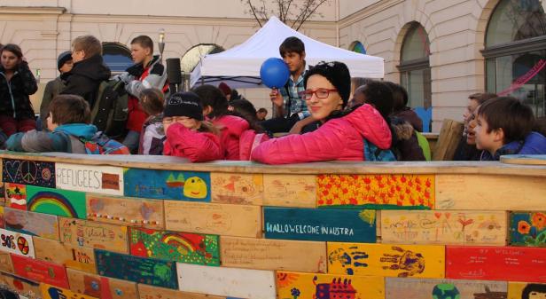 Eine Gruppe von Kindern steht hinter einer bemalten Holzwand mit der Aufschrift „Refugees Welcome“.