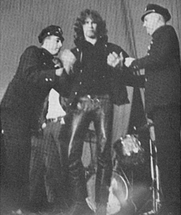 Zwei Polizisten führen Jim Morrison ab, der Lederhosen trägt.