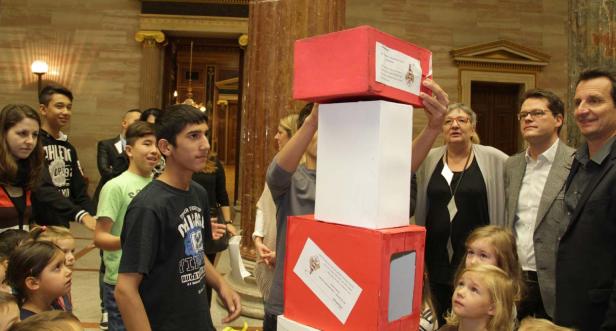 Eine Gruppe von Kindern und Erwachsenen betrachtet einen Turm aus roten und weißen Kisten.
