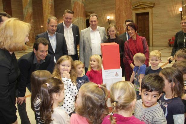 Eine Gruppe von Kindern und Erwachsenen steht um einen roten Kasten in einem Gebäude mit Säulen.