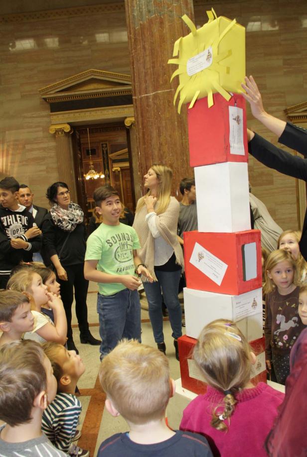 Eine Gruppe Kinder betrachtet einen Turm aus rot-weißen Kisten mit einer gelben Sonne an der Spitze.