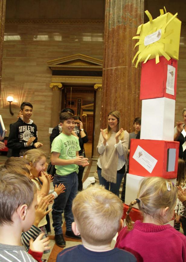 Eine Gruppe Kinder und Erwachsene steht um einen Turm aus roten, weißen und gelben Kartons und klatscht.