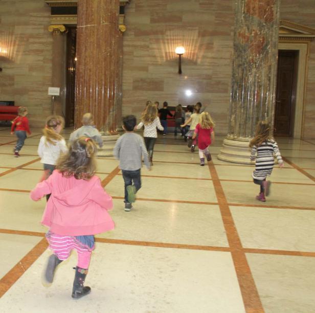 Eine Gruppe Kinder rennt in einem großen Saal mit Marmorsäulen.