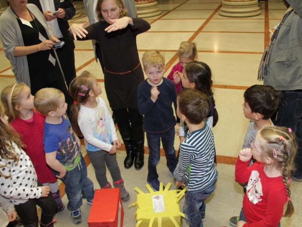 Eine Gruppe von Kindern steht um einen Tisch mit einer gelben Dekoration, während eine Frau gestikuliert.