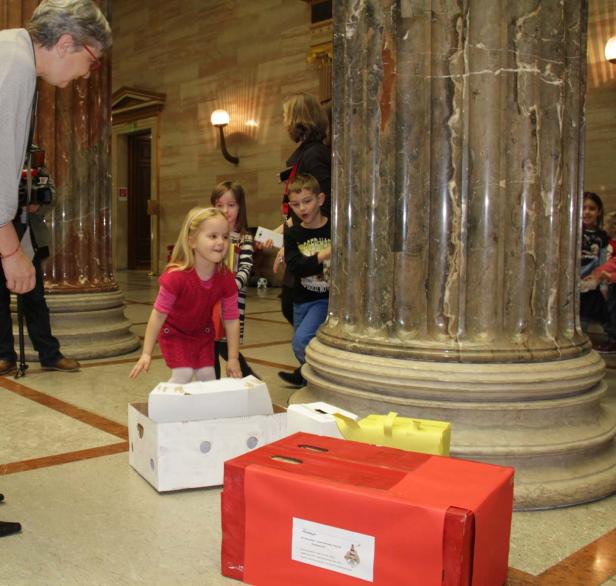 Kinder spielen mit bemalten Kartons in einer Halle mit Marmorsäulen.
