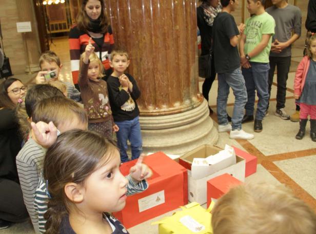 Eine Gruppe von Kindern steht um rote und gelbe Kisten herum und zeigt mit den Fingern.