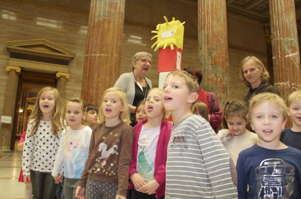Eine Gruppe von Kindern singt in einem Raum mit Säulen und einer Betreuerin.