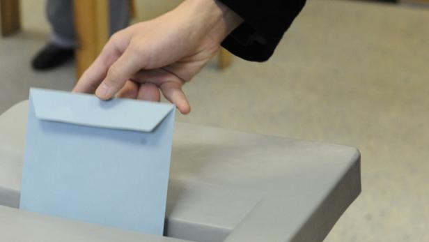 Eine Hand wirft einen hellblauen Briefumschlag in eine Wahlurne.