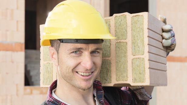 Ein Bauarbeiter mit gelbem Helm trägt einen isolierten Ziegelstein auf der Schulter.