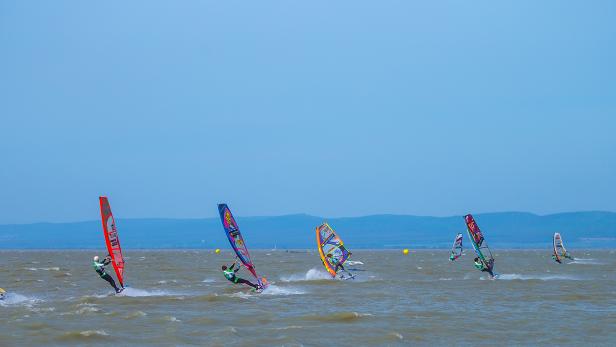 Mehrere Windsurfer rasen über einen See vor einer Hügellandschaft.