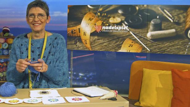 Eine Frau mit Brille strickt mit blauem Garn an einem Tisch voller Handarbeiten.