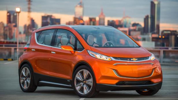 Ein orangefarbener Chevrolet Bolt steht vor einer Skyline bei Sonnenuntergang.