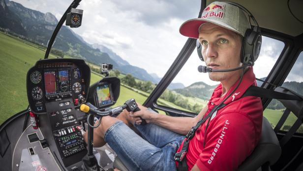 Ein Pilot mit Red Bull-Kappe steuert einen Hubschrauber über eine grüne Landschaft.