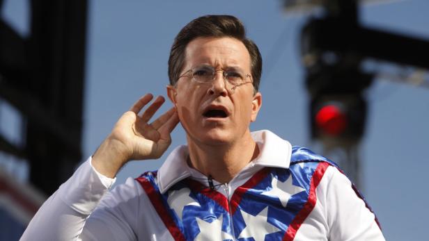 Stephen Colbert in einem weiß-blau-roten Anzug hält seine Hand ans Ohr.