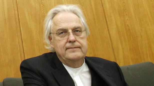 Ein älterer Mann mit weißem Haar und Brille sitzt vor einer Holzwand.