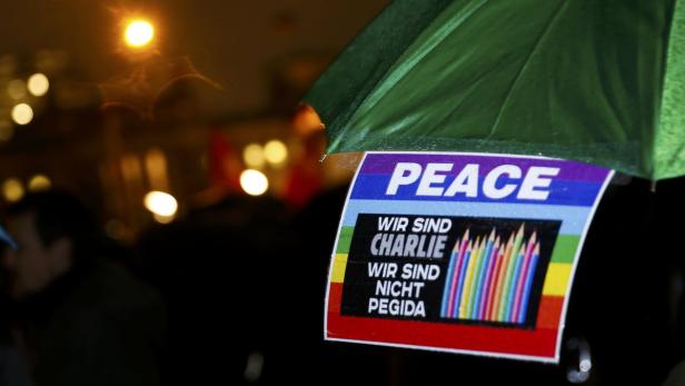 Ein Schild mit der Aufschrift „Peace, Wir sind Charlie, Wir sind nicht Pegida“ unter einem grünen Regenschirm.