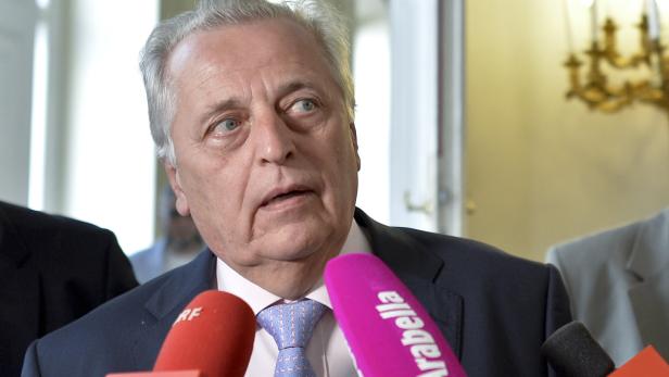 Ein älterer Mann spricht in mehrere Mikrofone vor einem hellen Hintergrund.