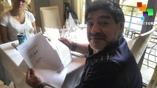 Diego Maradona sitzt an einem Tisch und hält ein signiertes Dokument in der Hand.