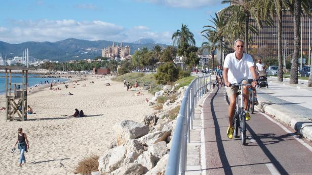 Ein Mann fährt mit dem Fahrrad auf einer Promenade entlang eines Strandes mit Badegästen und der Kathedrale von Palma im Hintergrund.