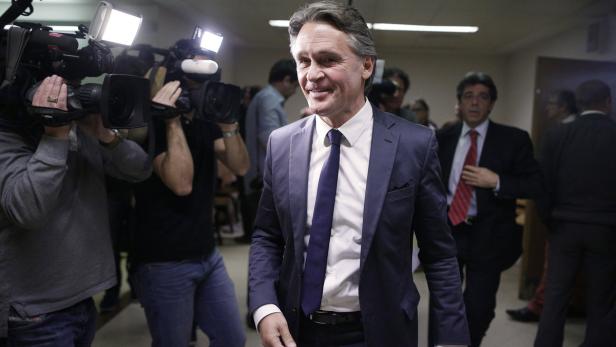 Ein Mann in Anzug lächelt, umgeben von Kameraleuten.