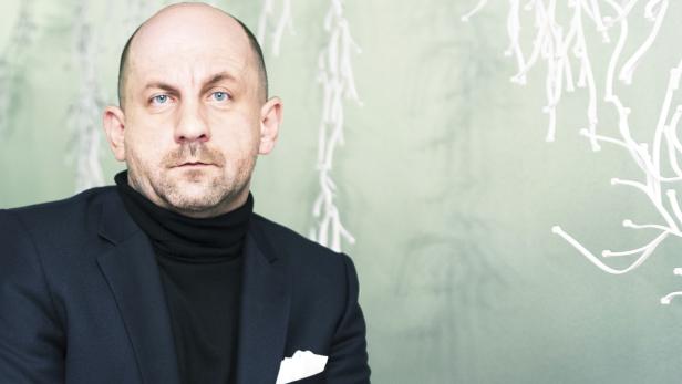 Ein Mann mit blauen Augen, einem schwarzen Rollkragenpullover und einem dunklen Sakko blickt in die Kamera.