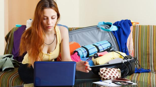 Eine Frau sitzt vor einem offenen Koffer und plant eine Reise am Laptop.