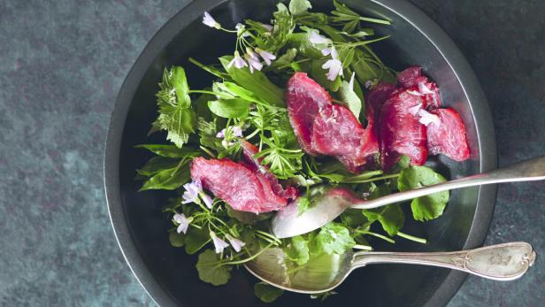 Ein Salat mit Wildkräutern, kleinen lila Blüten und Scheiben Räucherfleisch wird mit zwei Löffeln serviert.