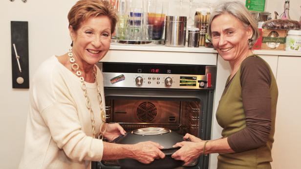Zwei Frauen hantieren mit einem Topf vor einem geöffneten Backofen.