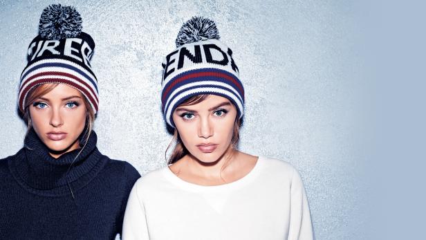 Zwei Frauen posieren mit gestreiften Mützen mit Bommeln vor einem hellblauen Hintergrund.