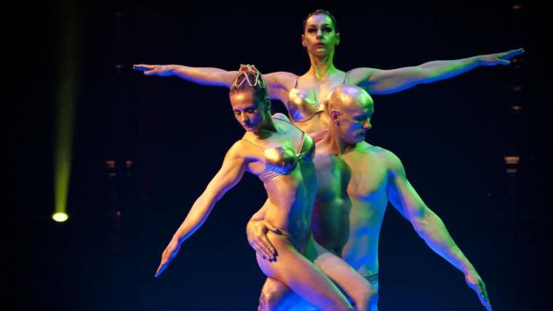 Drei Akrobaten in goldfarbenen Kostümen posieren auf einer Bühne.