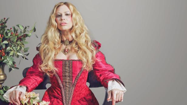 Eine Frau in einem roten Renaissance-Kleid mit goldenem Schmuck sitzt auf einem Stuhl.