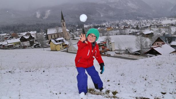 Ein Kind mit Brille wirft einen Schneeball vor einer verschneiten Landschaft mit Kirche.