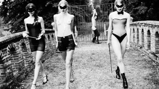 Drei Frauen mit Masken und Peitschen gehen auf einer Brücke entlang.