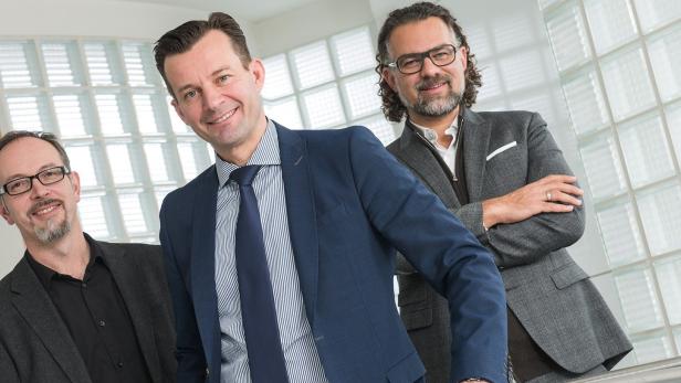 Drei Männer in Anzügen posieren vor einer Wand aus Glasbausteinen.