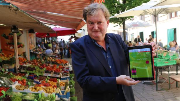 Ein Mann präsentiert ein Tablet auf einem Marktstand mit frischem Obst und Gemüse.