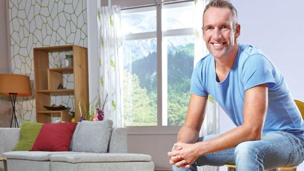 Ein lächelnder Mann sitzt in einem hellen Wohnzimmer vor einem Fenster mit Bergblick.