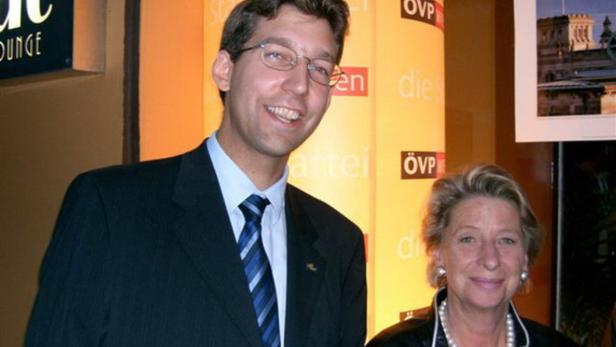 Ein Mann im Anzug und eine Frau mit Perlenkette posieren vor einem ÖVP-Hintergrund.