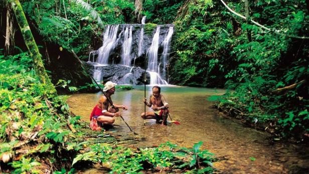 Zwei indigene Männer hocken in einem Bach vor einem Wasserfall im Dschungel.