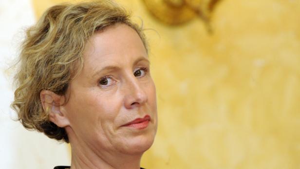 Porträt einer Frau mit blond gelocktem Haar vor einem gelben Hintergrund.
