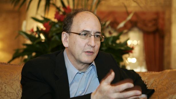 Ein Mann mit Brille gestikuliert vor einer Zimmerpflanze.