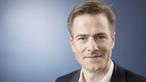 Ein lächelnder Mann mit blonden Haaren und einem dunklen Anzug vor blauem Hintergrund.