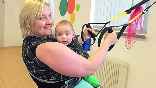 Eine Frau hält ein Baby in einer Trage und trainiert mit einem TRX-Band.