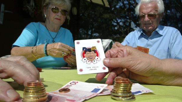 Zwei ältere Menschen spielen Karten um Geld, ein Joker liegt auf dem Tisch.