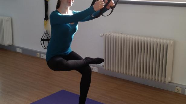 Eine Frau macht Kniebeugen mit einem gelben TRX-Band auf einer lila Matte.
