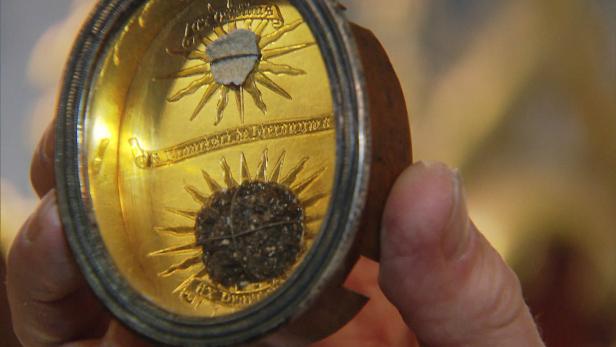 Eine Hand hält eine goldene Dose mit zwei stilisierten Sonnen und Reliquien darin.