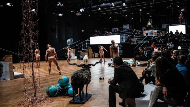 Eine Theaterszene mit Darstellern, einem Schaf und Zuschauern.