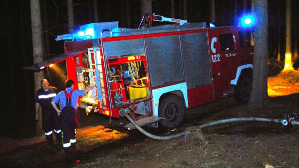 Zwei Feuerwehrleute stehen nachts neben einem roten Feuerwehrwagen im Wald.