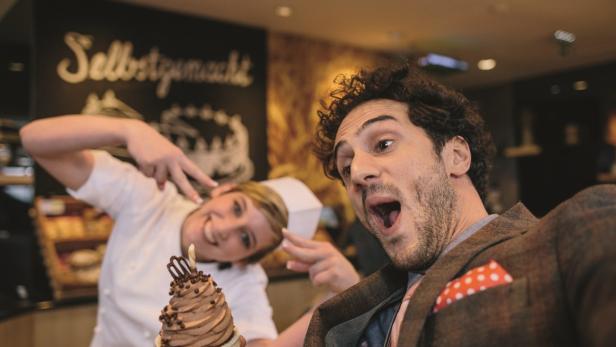 Ein Mann macht ein Selfie mit einem großen Schokoladenmuffin, während eine Bäckerin im Hintergrund posiert.