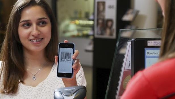 Eine junge Frau zeigt ein Smartphone mit einem Barcode an einer Kasse vor.