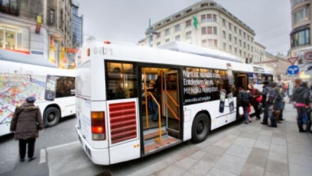 Ein weißer Bus hält an einer Haltestelle in der Stadt, Fahrgäste steigen ein und aus.